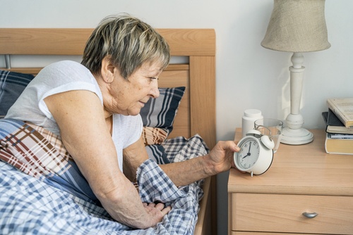 An elderly woman sitting up in bed and reaching for an alarm clock on her nightstand.