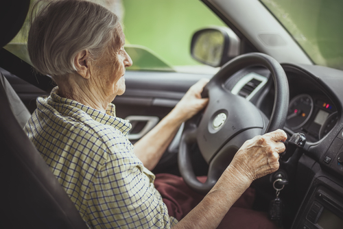 An elderly woman wearing a plaid shirt holding the steering wheel while driving a car.