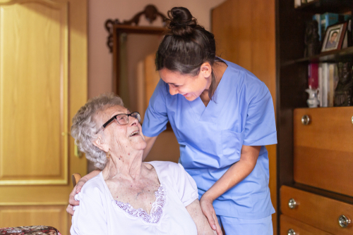 A smiling caregiver in blue scrubs leaning down to speak with an elderly woman seated in her home.