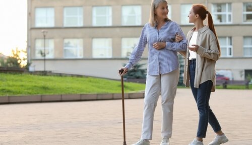 A young woman supporting an elderly woman with a cane who may be managing osteoporosis, walking together outdoors.