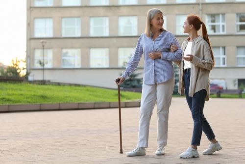 A young woman supporting an elderly woman with a cane who may be managing osteoporosis, walking together outdoors.