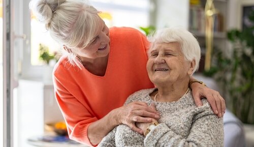 An adult daughter smiling and supporting her elderly mother at home.