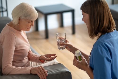 Caregiver handing a glass of water and medication bottle to an elderly woman holding pills in her hand