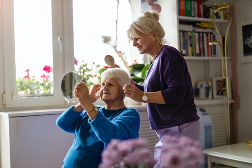 Smiling female caregiver assisting an elderly woman with styling her white hair while she looks into a small hand mirror.