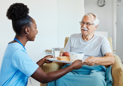 Young female caregiver serving a breakfast tray to a smiling senior man seated in an armchair.