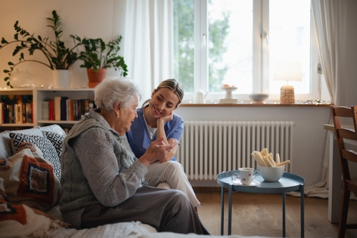 A smiling female care worker sitting on a sofa and conversing with an elderly woman in a cosy living room.